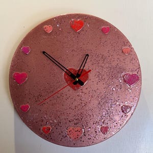 May include: A circular wall clock with a rose gold glitter finish. The clock face features twelve small heart-shaped markers in various shades of pink and red. A larger red heart sits at the centre, with two black hour and minute hands and a thin red second hand.