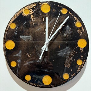 May include: A circular wall clock with a glossy black face featuring gold-coloured circular hour markers. The surface has a marbled effect with metallic copper and white accents. Three plain white clock hands are centred on the clock face.