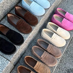 May include: A collection of suede loafers in various colors, including light blue, brown, black, pink, beige, and tan. The shoes are arranged on a set of gray stone steps, showcasing the different color options available.
