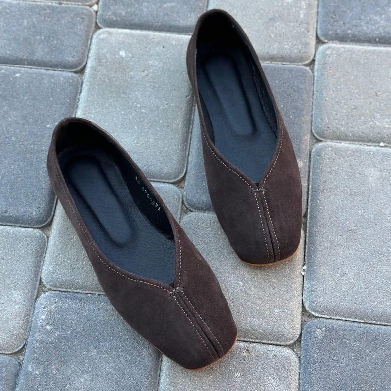 Pode incluir: Um par de sapatilhas de camur&ccedil;a castanhas escuras. Os sapatos t&ecirc;m um design de bico quadrado e uma costura costurada no centro. As solas s&atilde;o pretas e os sapatos s&atilde;o exibidos numa superf&iacute;cie de pedra cinzenta.