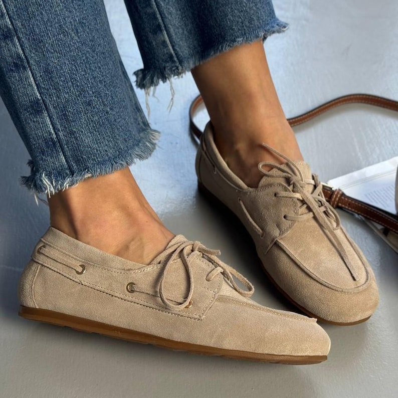 May include: Beige suede boat shoes with a lace-up design and a brown sole. The shoes have a classic, casual style, perfect for everyday wear. The shoes are shown with blue jeans.