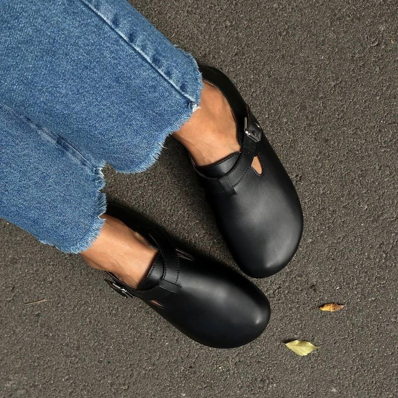May include: Black leather clogs with a buckle closure and a strap across the top. The clogs are worn with blue jeans. The shoes are a classic style, suitable for everyday wear.