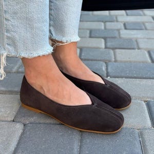 Pode incluir: Um par de sapatilhas de camur&ccedil;a castanho escuro com bico redondo e salto baixo. Os sapatos t&ecirc;m um design costurado na parte superior e uma sola castanho claro. As sapatilhas s&atilde;o usadas com jeans azul claro.