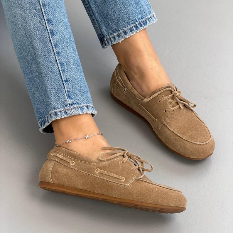 May include: Tan suede boat shoes with laces. The shoes are worn with blue jeans. The shoes have a brown sole and a light brown suede upper. A silver anklet with small charms is visible on the ankle.