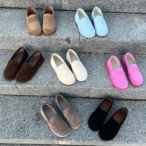 May include: A collection of suede loafers in various colors, including brown, light blue, dark brown, white, pink, taupe, and black. The shoes are arranged on a set of gray stone steps, showcasing their different hues and styles.