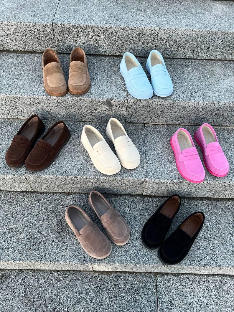 May include: A collection of suede loafers in various colors, including brown, light blue, dark brown, white, pink, taupe, and black. The shoes are arranged on a set of gray stone steps, showcasing their different hues and styles.