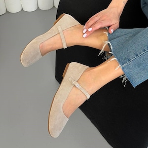 May include: Beige suede Mary Jane flats with a rounded toe and a delicate ankle strap. The shoes have a low heel and a neutral color, suitable for various outfits. The flats are a stylish and comfortable footwear option.