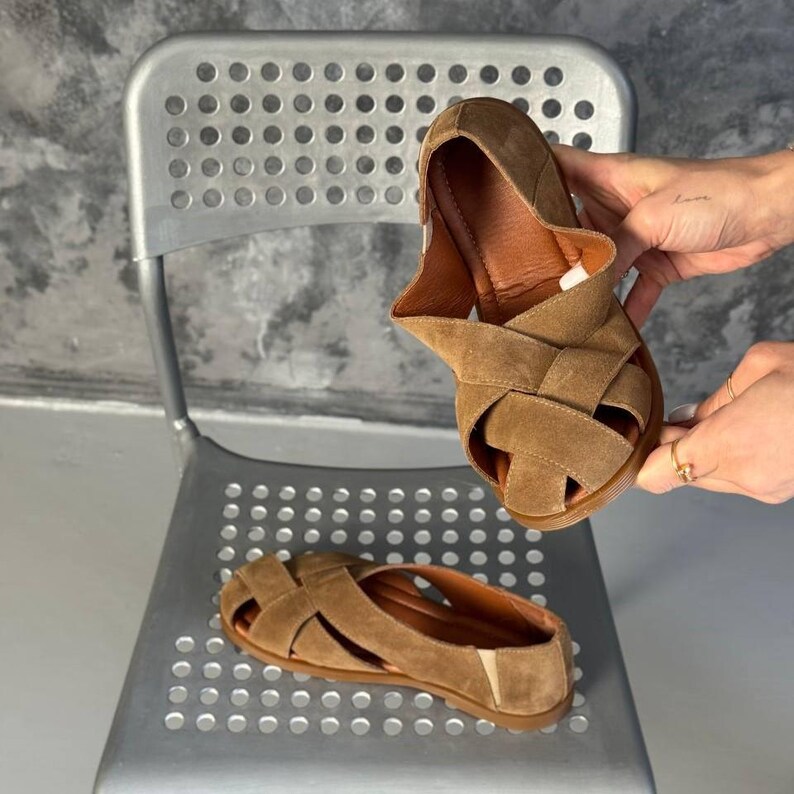 May include: A pair of brown suede sandals with a woven strap design. The sandals have a flat sole and a leather interior. One sandal is held up by a person's hands, while the other rests on a metal chair.