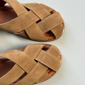 May include: A pair of tan suede sandals with a woven strap design. The sandals have a flat sole and a light brown interior. The straps are stitched together, creating a crisscross pattern across the top of the foot.
