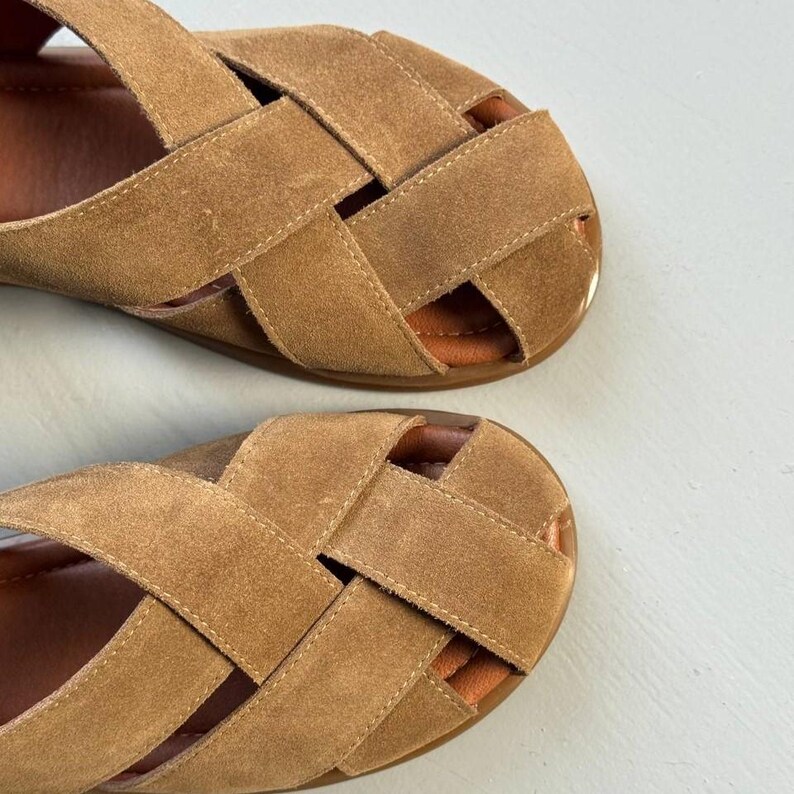 May include: A pair of tan suede sandals with a woven strap design. The sandals have a flat sole and a light brown interior. The straps are stitched together, creating a crisscross pattern across the top of the foot.
