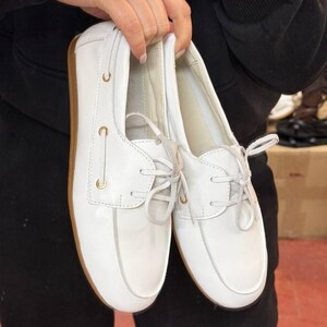 May include: A pair of white leather boat shoes with light brown soles. The shoes have laces and gold-colored eyelets. The shoes are held up by a person.