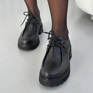 May include: Black leather shoes with a chunky sole and lace-up closure. The shoes have a rounded toe and a thick, textured sole. The laces are black and tied in a bow. The shoes are worn with black sheer tights.