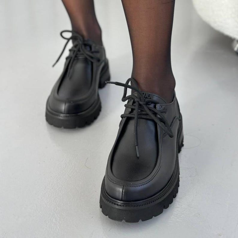 May include: Black leather shoes with a chunky sole and lace-up closure. The shoes have a rounded toe and a thick, textured sole. The laces are black and tied in a bow. The shoes are worn with black sheer tights.