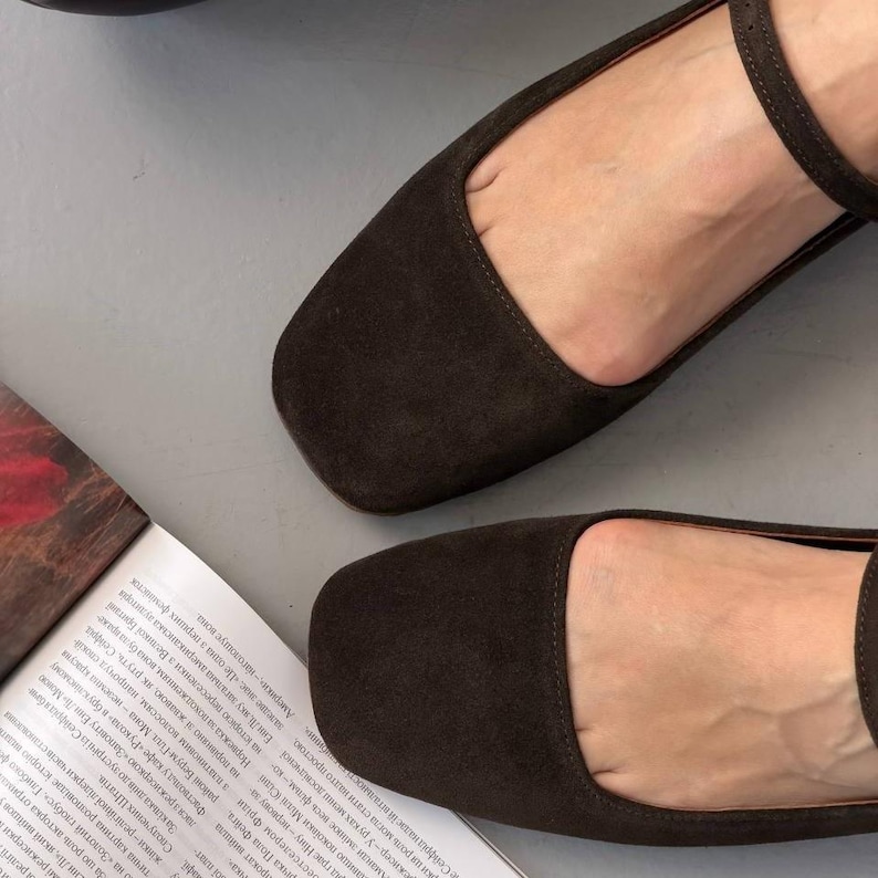 May include: Dark brown suede flats with a square toe and ankle strap. The shoes are displayed on a light gray surface, with a magazine visible in the background. These flats are a stylish choice for everyday wear.