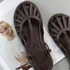 Puede incluir: Sandalias de cuero marr&oacute;n oscuro con dise&ntilde;o de tira en T y cierre de hebilla. Las sandalias presentan una parte superior en forma de jaula con m&uacute;ltiples correas y suela plana. Los zapatos se muestran en una p&aacute;gina de revista.