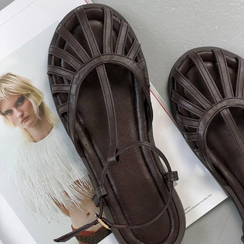 Puede incluir: Sandalias de cuero marr&oacute;n oscuro con dise&ntilde;o de tira en T y cierre de hebilla. Las sandalias presentan una parte superior en forma de jaula con m&uacute;ltiples correas y suela plana. Los zapatos se muestran en una p&aacute;gina de revista.