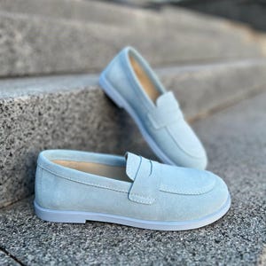 May include: Two pairs of light blue suede loafers. One pair is in the foreground, with the other pair slightly behind and above. The shoes have a classic penny loafer design with a rounded toe and a low heel. The shoes are on a gray stone surface.
