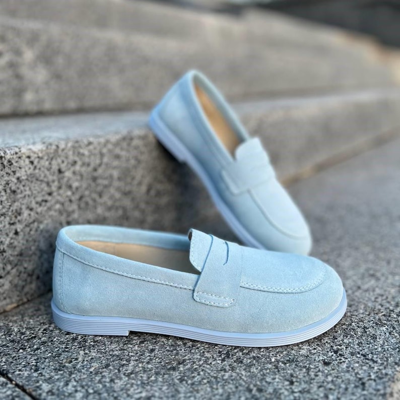 May include: Two pairs of light blue suede loafers. One pair is in the foreground, with the other pair slightly behind and above. The shoes have a classic penny loafer design with a rounded toe and a low heel. The shoes are on a gray stone surface.