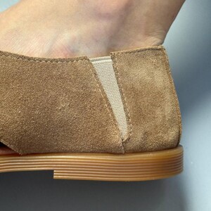 May include: Close-up of a brown suede shoe with a tan elastic panel. The shoe has a rounded toe and a flat, light brown sole. The suede has a soft, textured appearance.