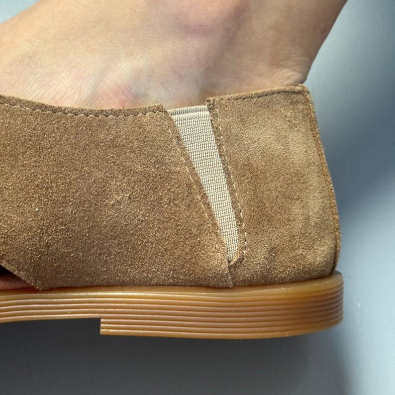 May include: Close-up of a brown suede shoe with a tan elastic panel. The shoe has a rounded toe and a flat, light brown sole. The suede has a soft, textured appearance.