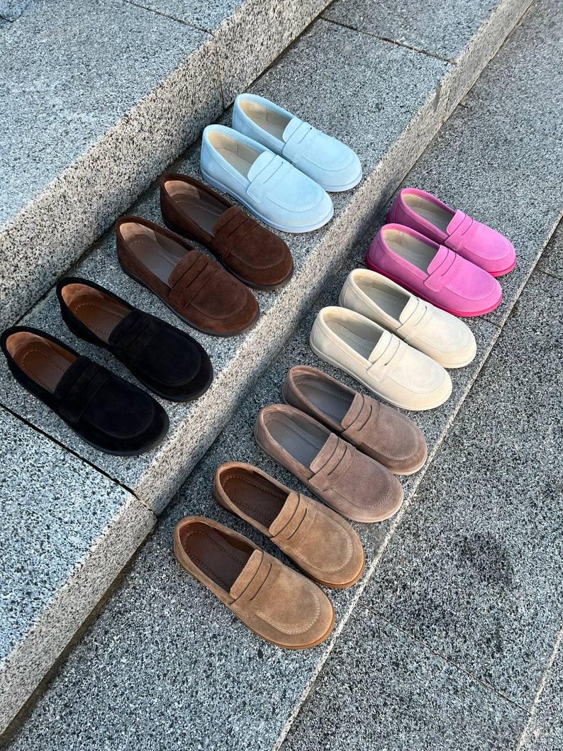 May include: A collection of suede loafers in various colors, including light blue, brown, black, pink, beige, and tan. The shoes are arranged on a set of gray stone steps, showcasing the different color options available.
