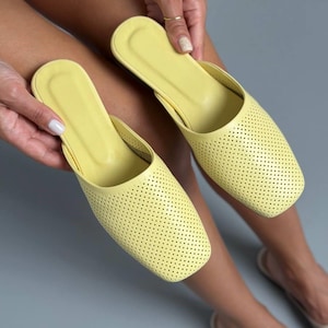 May include: A pair of light yellow slip-on mules featuring a square toe design and a perforated upper surface. The shoes have a smooth, solid-colored interior footbed and a consistent, uniform texture across the exterior of the footwear.