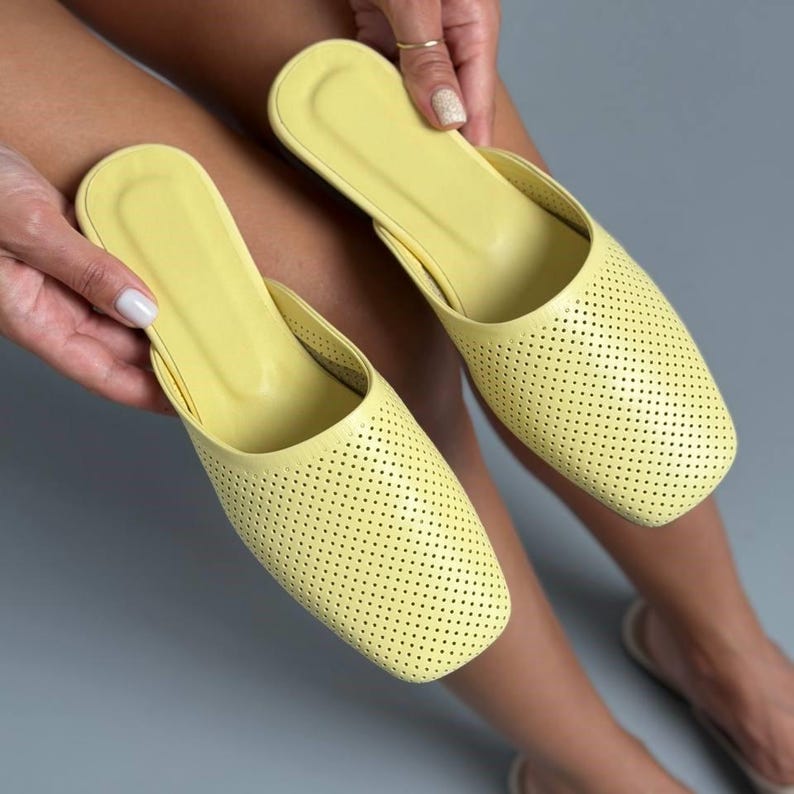 May include: A pair of light yellow slip-on mules featuring a square toe design and a perforated upper surface. The shoes have a smooth, solid-colored interior footbed and a consistent, uniform texture across the exterior of the footwear.