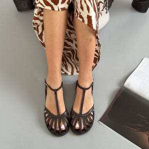 Puede incluir: Sandalias de cuero marr&oacute;n oscuro con dise&ntilde;o de correa en T y cierre de hebilla. Las sandalias tienen una estructura similar a una jaula alrededor del pie. La persona lleva una falda con estampado de cebra.
