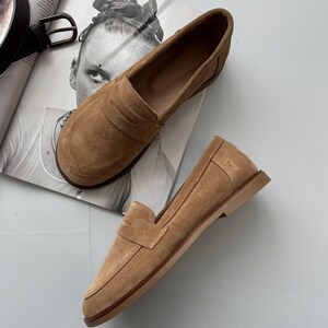 May include: A pair of tan suede penny loafers featuring a classic moc-toe construction, a horizontal strap across the vamp, and a flat, low-profile sole. The shoes have a soft, brushed texture and a rounded toe design with visible stitching details.