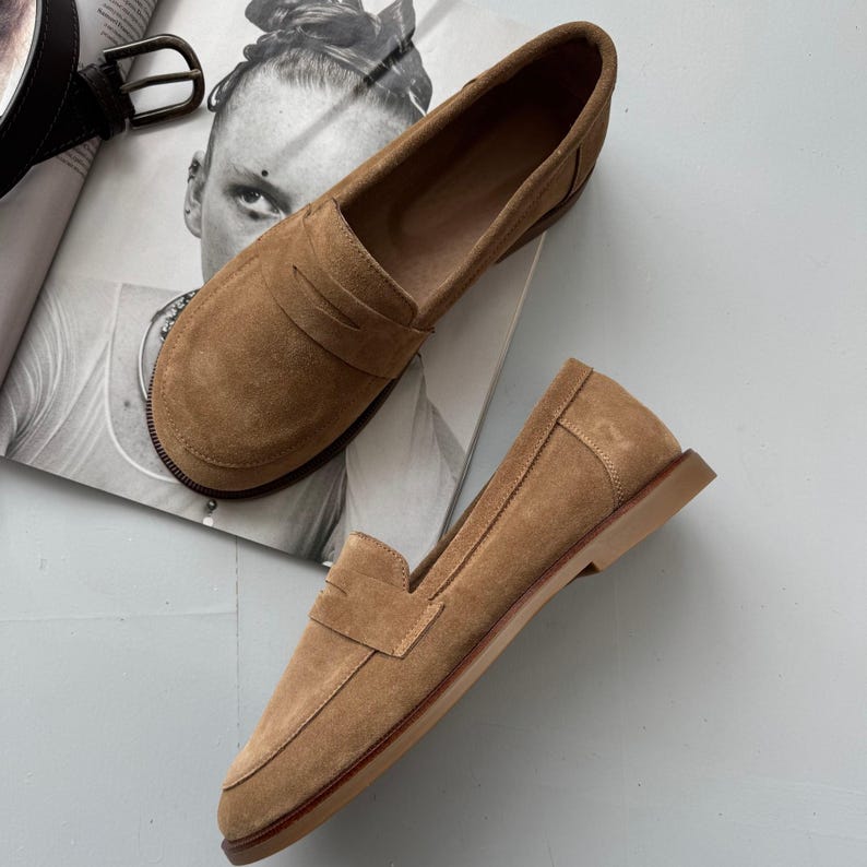 May include: A pair of tan suede penny loafers featuring a classic moc-toe construction, a horizontal strap across the vamp, and a flat, low-profile sole. The shoes have a soft, brushed texture and a rounded toe design with visible stitching details.