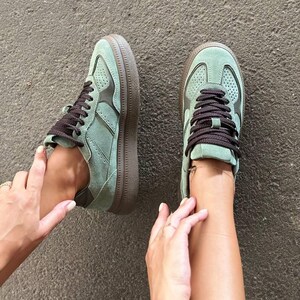 May include: A pair of sage green suede sneakers featuring dark brown laces, dark brown trim accents, and a thick dark brown rubber sole. The upper portion of the sneakers includes perforated detailing on the toe box and side panels for texture.