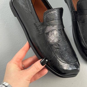 May include: Black leather loafers with a glossy finish. The shoes have a classic design with a rounded toe and a strap detail across the top. The soles are black and the interior is a light brown color.