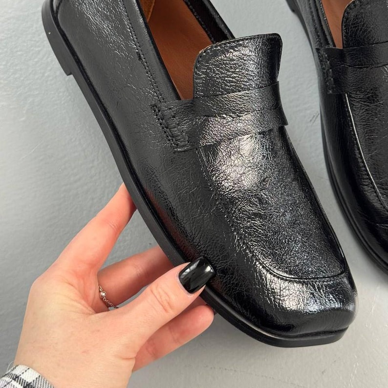 May include: Black leather loafers with a glossy finish. The shoes have a classic design with a rounded toe and a strap detail across the top. The soles are black and the interior is a light brown color.