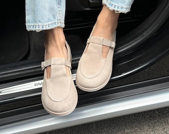May include: A pair of beige suede T-strap shoes featuring a rounded toe, a flat sole, and a thin adjustable strap across the instep with a small silver-toned metal buckle. The shoes have a smooth, matte texture and a simple, clean design.