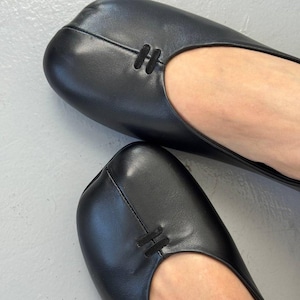 Op de afbeelding: Zwarte leren ballerina's met een vierkante neus. De schoenen hebben een eenvoudig, elegant ontwerp, met een klein decoratief detail aan de bovenkant. De afbeelding toont een close-up van het schoeisel.