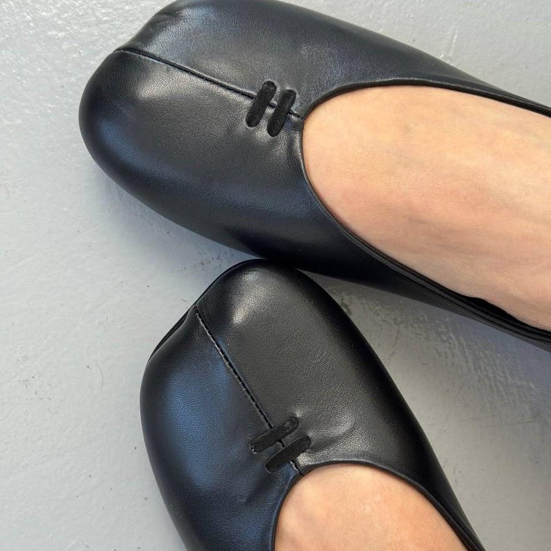 Op de afbeelding: Zwarte leren ballerina's met een vierkante neus. De schoenen hebben een eenvoudig, elegant ontwerp, met een klein decoratief detail aan de bovenkant. De afbeelding toont een close-up van het schoeisel.