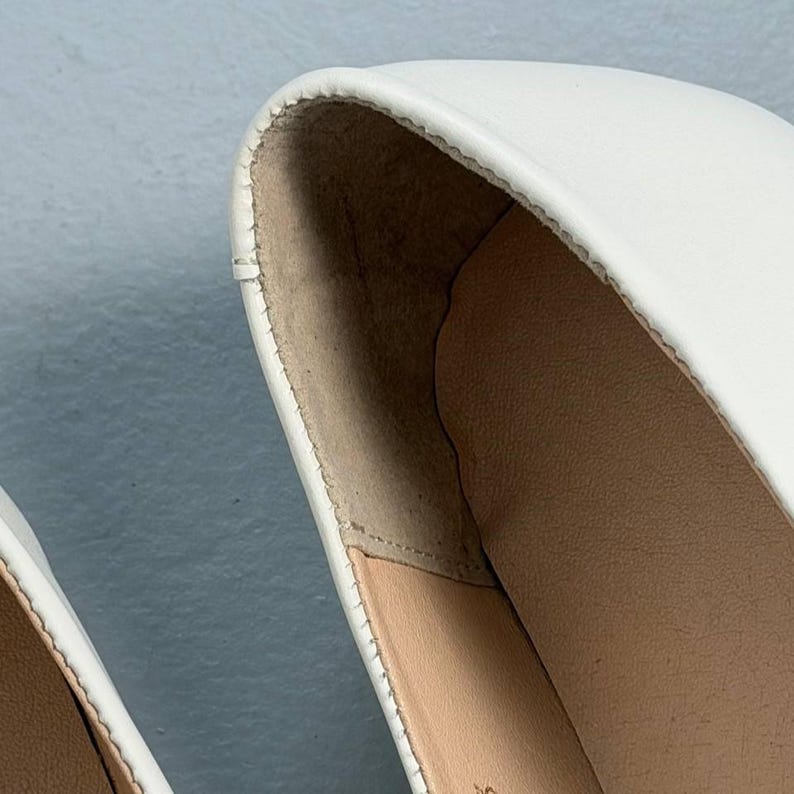 Op de afbeelding: Close-up van een witte schoen, met de gladde leren bovenkant en de stiksels langs de rand. De binnenkant van de schoen is lichtbruin van kleur. De schoen lijkt nieuw en ongedragen.
