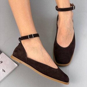 May include: Dark brown suede flats with ankle straps and small silver buckles. The shoes have a square toe design and a light brown sole. The flats are a stylish and comfortable footwear option.