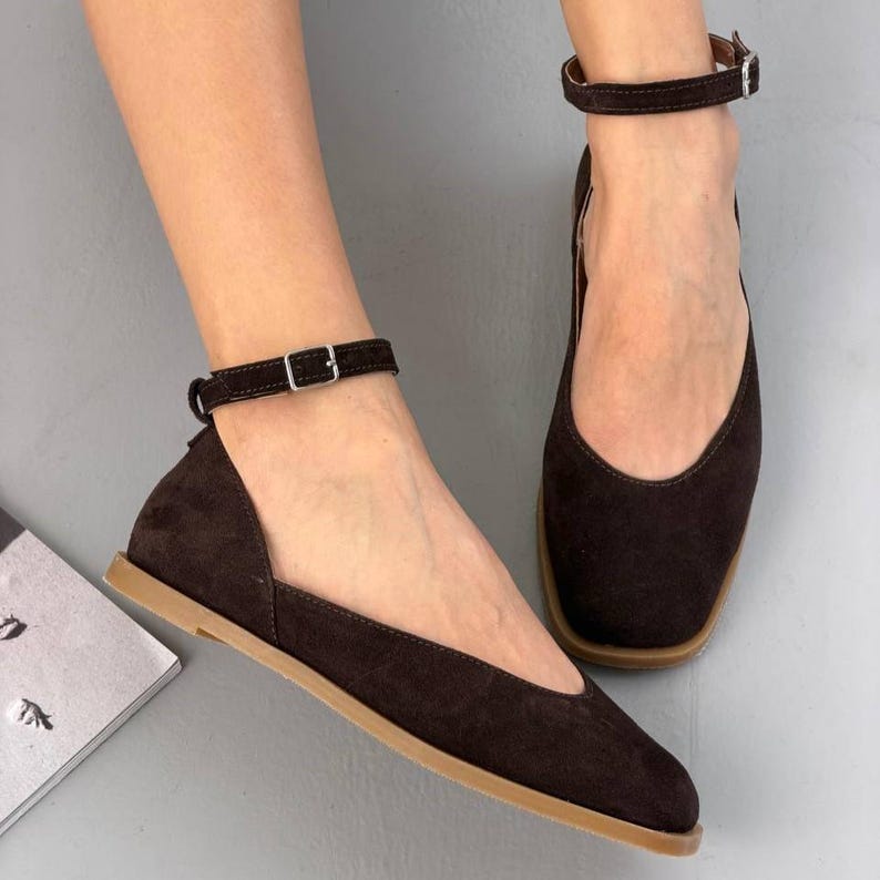 May include: Dark brown suede flats with ankle straps and small silver buckles. The shoes have a square toe design and a light brown sole. The flats are a stylish and comfortable footwear option.