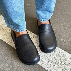 May include: A pair of black leather penny loafers. The shoes have a rounded toe and a strap across the top. The shoes are worn with blue jeans. The shoes are on a gray surface with a white stripe.