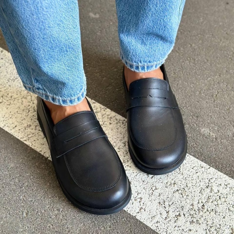 May include: A pair of black leather penny loafers. The shoes have a rounded toe and a strap across the top. The shoes are worn with blue jeans. The shoes are on a gray surface with a white stripe.