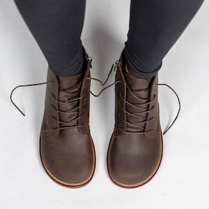 Puede incluir: Un par de botines de cuero marrón oscuro con punta redondeada, cordones frontales a juego y cierre de cremallera lateral. Las botas presentan costuras visibles a lo largo de la suela y se llevan puestas con unos leggings de color negro.