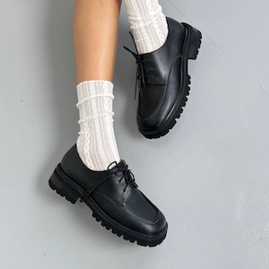 May include: Black leather Oxford shoes with a square toe and thick lug sole. The shoes have a lace-up closure and are paired with white lace socks. The shoes are shown against a gray background.