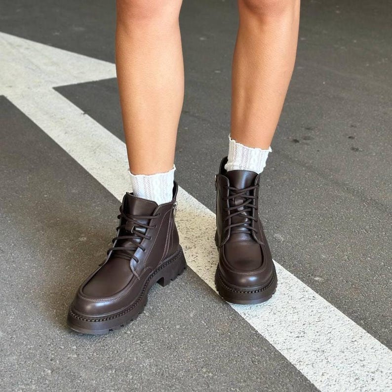 May include: A pair of brown leather combat boots with a lace-up design and a chunky sole. The boots have a side zipper and are worn with white ribbed socks. The boots are on a gray surface with a white stripe.