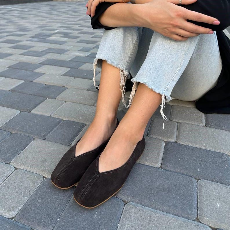 Pode incluir: Um par de sapatilhas de camur&ccedil;a castanhas escuras com bico quadrado e decote em V. Os sapatos t&ecirc;m um design minimalista e uma paleta de cores neutras. As solas s&atilde;o de cor castanho claro.