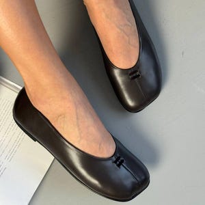 Op de afbeelding: Bruine leren ballerina's met een vierkante neus en een klein decoratief detail op de wreef. De schoenen hebben een gladde, gepolijste afwerking en een lage hak. De ballerina's zijn ontworpen voor comfort en stijl.