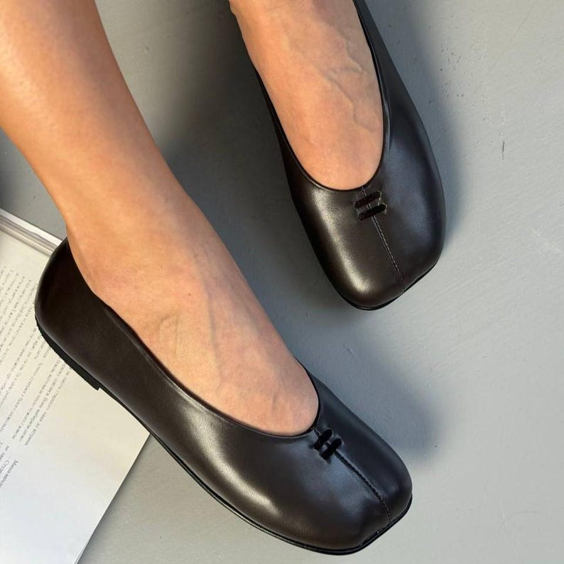 Op de afbeelding: Bruine leren ballerina's met een vierkante neus en een klein decoratief detail op de wreef. De schoenen hebben een gladde, gepolijste afwerking en een lage hak. De ballerina's zijn ontworpen voor comfort en stijl.