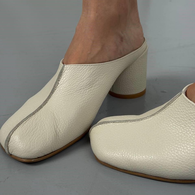 May include: Cream-colored leather mules with a square toe and a block heel. The shoes have a textured surface and a light gray seam detail. The heel height is approximately 3 inches.