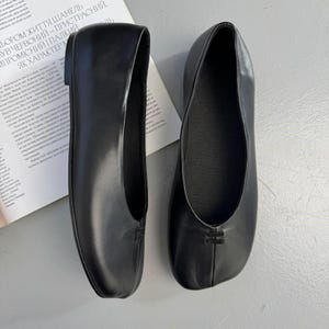 Op de afbeelding: Een paar zwarte leren ballerina's. De schoenen hebben een ronde neus en een klein decoratief detail op de wreef. De ballerina's worden gepresenteerd op een lichtgrijze ondergrond.