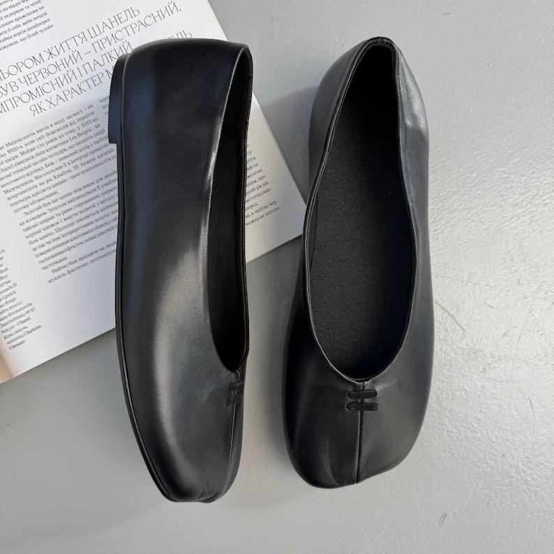 Op de afbeelding: Een paar zwarte leren ballerina's. De schoenen hebben een ronde neus en een klein decoratief detail op de wreef. De ballerina's worden gepresenteerd op een lichtgrijze ondergrond.